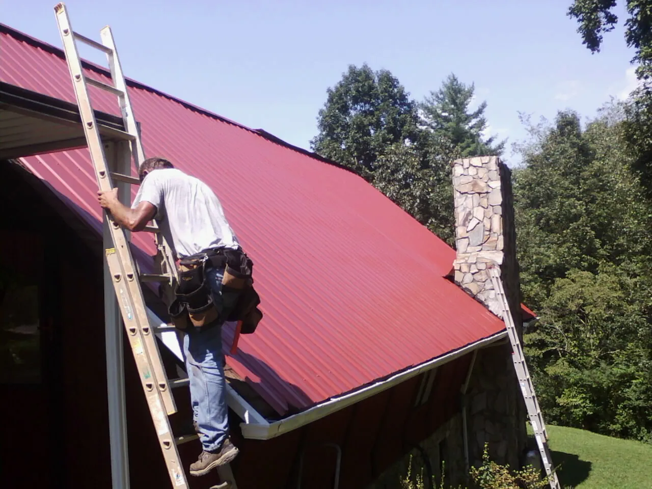 Roofer on a ladder beside a red metal roof and stone chimney — Soffit & Fascia Repair by Roofing Brothers in Burr Oak